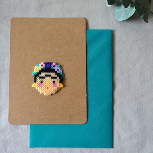 Puede incluir: Una tarjeta de felicitación hecha a mano con una base de cartulina marrón, un sobre turquesa y un retrato de arte de cuentas de colores de una mujer con una corona floral. El retrato está hecho de pequeñas cuentas de colores.