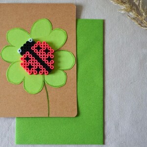 May include: A handmade greeting card with a red and black ladybird made of small beads, set on a green four-leaf clover. The card is on a brown background, with a green envelope and a sprig of dried grass.