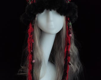 Puede incluir: Un sombrero artesanal con orejas de gato, con un diseño texturizado en rojo, negro y verde. El sombrero tiene un ribete de piel sintética negra y largas hebras rojas y negras con pequeños acentos florales rosas. En una cabeza de maniquí.
