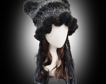 May include: A black and gray faux fur hat with ear-like protrusions and a fluffy brim. The hat is adorned with colorful flecks and is paired with long, fringed, black faux fur extensions. The hat is displayed on a mannequin head.