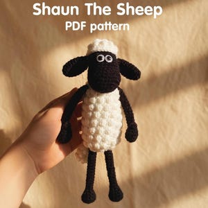 Könnte beinhalten: Gehäkelte Shaun das Schaf Puppe mit weißem Körper, schwarzem Kopf, Ohren, Armen und Beinen. Die Puppe hat weiße Augen und hält ein PDF-Muster. Der Text "Shaun The Sheep PDF pattern" befindet sich oben im Bild.