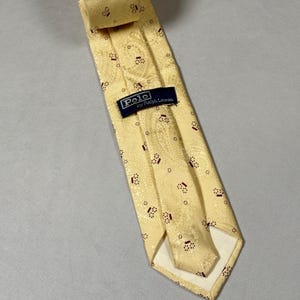 May include: A pale yellow necktie featuring a tonal paisley pattern and small dark red geometric accents. The tie includes a dark blue rectangular label stitched on the back that reads Polo by Ralph Lauren in white lettering.
