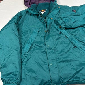 May include: A teal Columbia jacket featuring a high collar with a dark teal fleece lining, a front zipper closure with a snap-button placket, long sleeves with adjustable cuffs, and a small embroidered logo on the chest and sleeve.