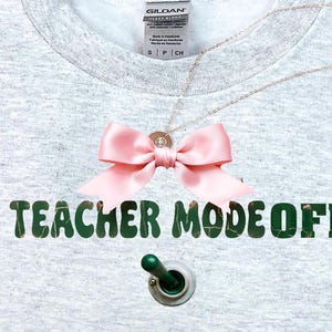 Puede incluir: Una sudadera gris claro con el texto "TEACHER MODE OFF" en verde. Un lazo rosa y un collar están prendidos a la sudadera. Un interruptor basculante verde está debajo del texto.