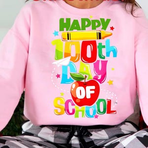 May include: Pink sweatshirt with a colorful graphic that reads "Happy 100th Day of School." The design includes a yellow crayon, a paper airplane, a red apple, and stars. The sweatshirt is a crew neck style.