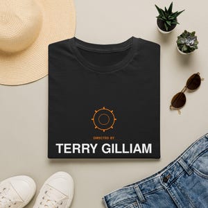 May include: A black short-sleeved t-shirt featuring an orange circular gear icon above the orange text "DIRECTED BY" and the white text "TERRY GILLIAM" centered on the chest. The shirt is displayed flat with a round neckline and short sleeves.
