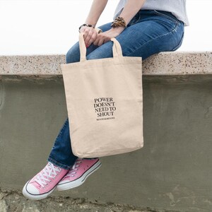 May include: A beige canvas tote bag with the text "POWER DOESN'T NEED TO SHOUT" in black. The bag is held by a person wearing blue jeans and pink sneakers.