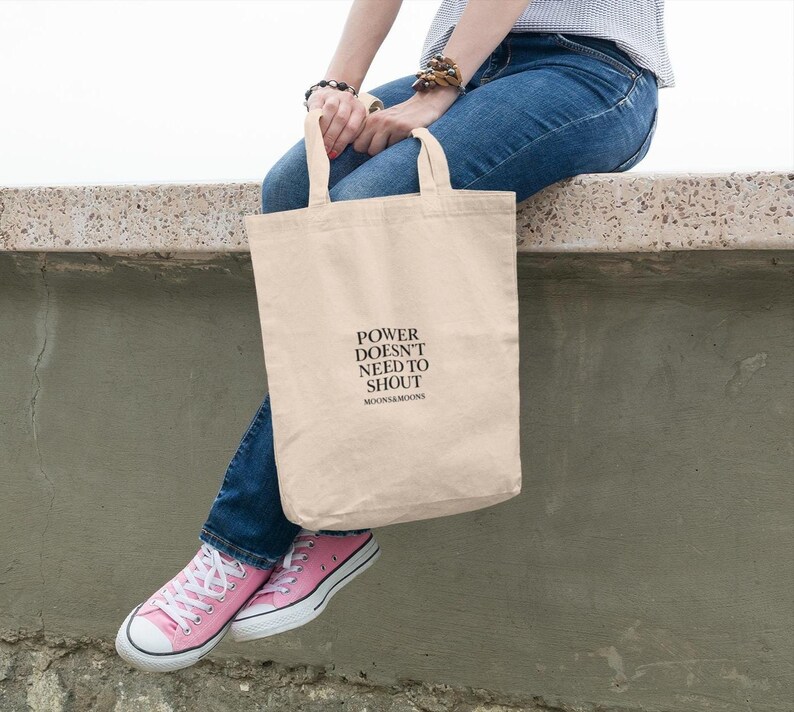 May include: A beige canvas tote bag with the text "POWER DOESN'T NEED TO SHOUT" in black. The bag is held by a person wearing blue jeans and pink sneakers.