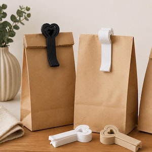 Puede incluir: Bolsas de papel marrón con clips para bolsas en forma de corazón en blanco y negro. Varios clips están apilados sobre una superficie de madera. Un jarrón con vegetación está en el fondo. Los clips están diseñados para sellar las bolsas.