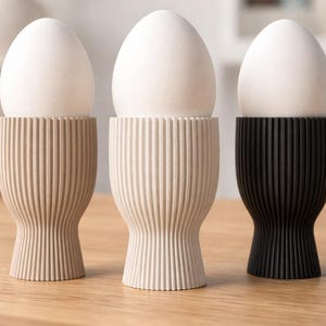 Könnte beinhalten: Drei Eierbecher, die jeweils ein weißes Ei halten. Die Eierbecher sind in drei Farben erhältlich: Beige, Off-White und Schwarz. Die Becher haben ein geripptes Design und einen ausgestellten Boden.