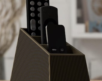 May include: A dark olive green remote control holder with a tiered, sloped design and vertical ribbed texture. The organizer contains three black remote controls of varying heights, with one remote featuring a visible menu button and icon.