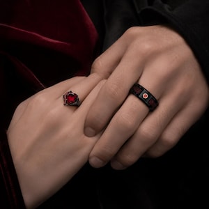 Puede incluir: Un anillo de metal negro con una piedra roja en forma de corazón y una banda negra con una incrustación circular roja. El anillo de corazón tiene pequeñas piedras rojas en el engaste, mientras que la banda muestra un punto rojo central.