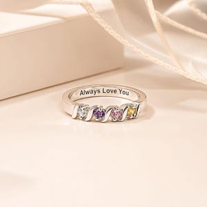 May include: A silver-toned ring featuring four round-cut stones in clear, purple, pink, and yellow colors set within a wavy metal band. The interior of the band is engraved with the text Always Love You in a clear, dark, sans-serif font.