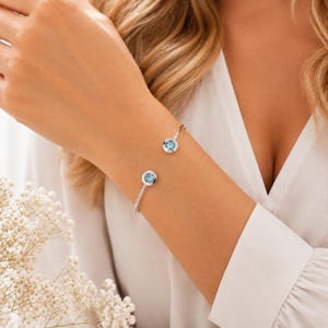 Op de afbeelding: Een zilverkleurige open armband met twee ronde zettingen, elk voorzien van een enkele ronde, lichtblauwe edelsteen. De armband heeft een gepolijste metalen afwerking en een eenvoudig, strak ontwerp dat om de pols rust.