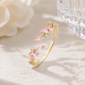 Pode incluir: Um anel aberto em tom dourado com dois motivos em forma de folha, adornados com pedras cor-de-rosa de corte marquise e pequenas pedras redondas transparentes. A banda é fina e polida, com as duas extremidades decorativas frente a frente.