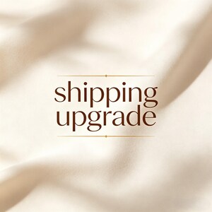 May include: A cream-coloured fabric background with soft folds and ripples. Centered in the image is the text "shipping upgrade" written in a dark brown serif font, framed by two thin horizontal lines with small diamond accents.