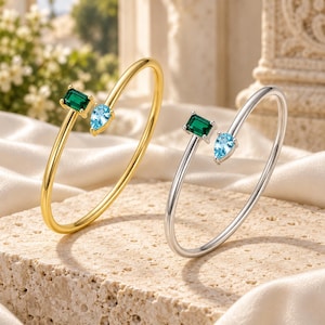 Op de afbeelding: Twee open armbanden, één in goudkleur en één in zilverkleur, elk voorzien van een rechthoekige smaragdgroene steen en een peervormige lichtblauwe steen aan de uiteinden. De armbanden hebben een dun, glad metalen ontwerp.