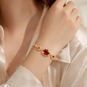 Puede incluir: Una pulsera de tono dorado con una gran piedra preciosa redonda de color rojo intenso en el centro, rodeada por un halo de pequeñas piedras claras. La cadena incluye dos piedras rojas redondas más pequeñas a cada lado, con engaste dorado.