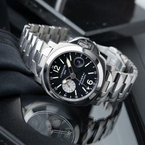 May include: A stainless steel Panerai Luminor GMT automatic watch with a black dial. The watch features luminous hands and markers, a date display, and a stainless steel bracelet. The watch is displayed on a reflective surface.