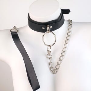 May include: Black leather choker with a large silver ring and chain detail. The choker features a strap that extends down the chest. The accessory is displayed on a white mannequin.