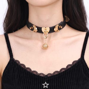 May include: A black leather choker necklace featuring gold-toned hardware, including two crescent moon accents and a central heart-shaped ring. A gold-toned filigree bell charm hangs from a lobster clasp attached to the heart ring.
