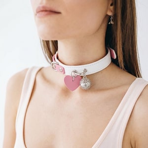 May include: White leather choker with pink accents and a pink heart pendant. The choker features a silver-colored bell charm and a buckle closure. The design is simple and elegant.