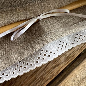 K&ouml;nnte beinhalten: Nahaufnahme eines Leinenstoffs mit wei&szlig;er Lochspitze. Ein Holzrundstab ist sichtbar, mit einem wei&szlig;en Satinband gebunden. Der Stoff hat eine nat&uuml;rliche beige Farbe, und die Spitze hat einen zarten, gewellten Rand. Der Hintergrund ist eine Holzoberfl&auml;che.