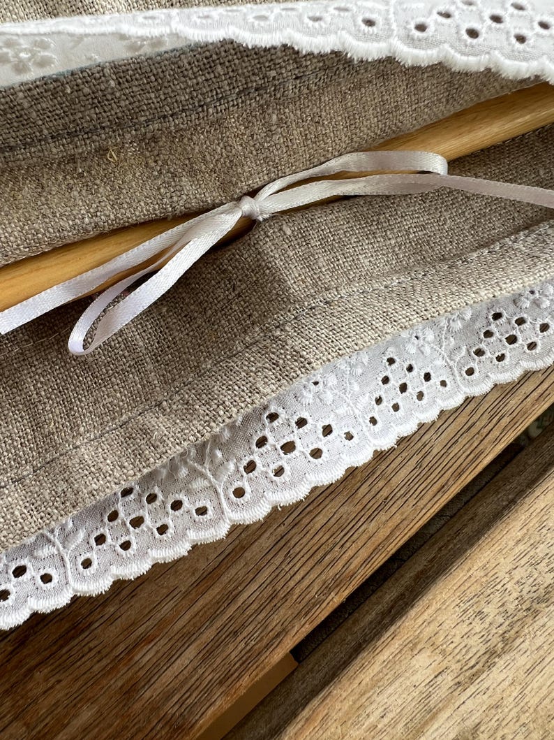 K&ouml;nnte beinhalten: Nahaufnahme eines Leinenstoffs mit wei&szlig;er Lochspitze. Ein Holzrundstab ist sichtbar, mit einem wei&szlig;en Satinband gebunden. Der Stoff hat eine nat&uuml;rliche beige Farbe, und die Spitze hat einen zarten, gewellten Rand. Der Hintergrund ist eine Holzoberfl&auml;che.