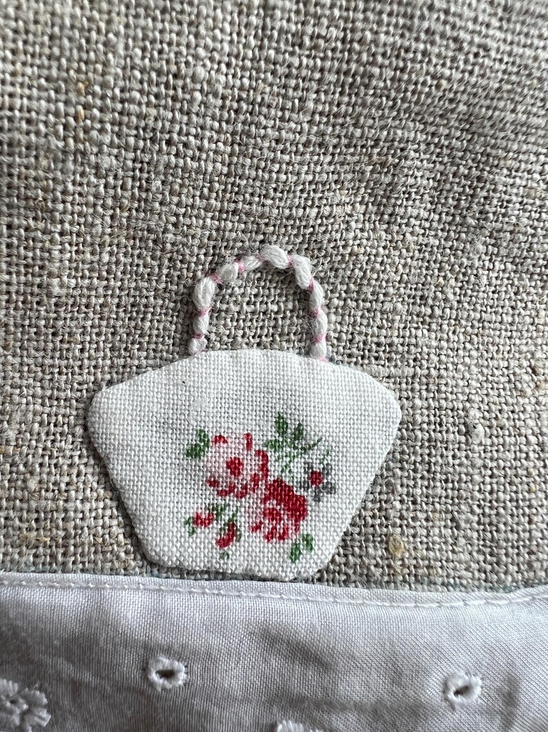 K&ouml;nnte beinhalten: Ein kleiner, wei&szlig;er Stofffleck in Form eines Korbes, bestickt mit roten und gr&uuml;nen Blumen. Der Korb hat einen rosa-wei&szlig;en Griff. Der Aufn&auml;her ist auf einen strukturierten, grauen Stoff gen&auml;ht.