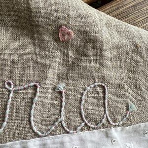 K&ouml;nnte beinhalten: Nahaufnahme eines Leinentextils mit gestickten Details. Das Wort "ria" ist in Schreibschrift mit hellblauem und rosa Garn gestickt. Ein kleines rosa Herz ist &uuml;ber den Buchstaben gestickt. Der untere Rand hat eine wei&szlig;e, floral gemusterte Bord&uuml;re.