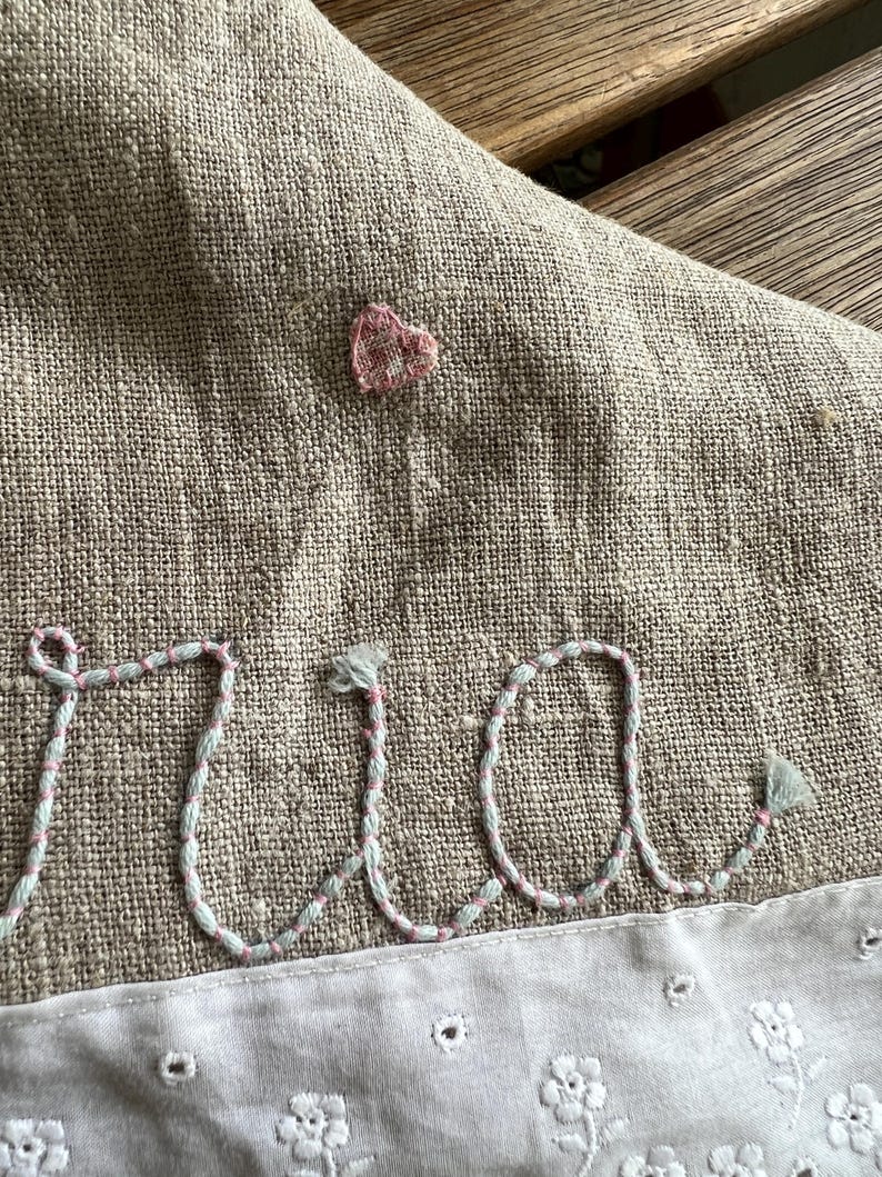 K&ouml;nnte beinhalten: Nahaufnahme eines Leinentextils mit gestickten Details. Das Wort "ria" ist in Schreibschrift mit hellblauem und rosa Garn gestickt. Ein kleines rosa Herz ist &uuml;ber den Buchstaben gestickt. Der untere Rand hat eine wei&szlig;e, floral gemusterte Bord&uuml;re.