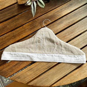 K&ouml;nnte beinhalten: Ein beiger Stoffkleiderb&uuml;gel mit dem Namen "Maria" in wei&szlig;er Schrift gestickt. Der untere Rand hat eine wei&szlig;e Spitzentrimmung. Eine kleine rosa Verzierung ist sichtbar.