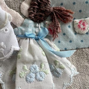 K&ouml;nnte beinhalten: Nahaufnahme von handgefertigten Stoffpuppen. Eine Puppe hat ein wei&szlig;es Kleid mit blauer Blumenstickerei und einem blauen Band. Eine andere Puppe hat ein hellblaues Kleid mit einer Tasche. Die Puppen haben braunes Garnhaar.