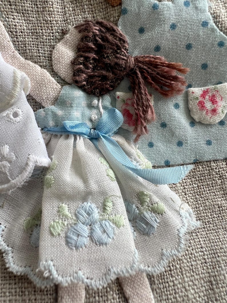 K&ouml;nnte beinhalten: Nahaufnahme von handgefertigten Stoffpuppen. Eine Puppe hat ein wei&szlig;es Kleid mit blauer Blumenstickerei und einem blauen Band. Eine andere Puppe hat ein hellblaues Kleid mit einer Tasche. Die Puppen haben braunes Garnhaar.