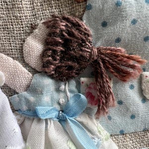 K&ouml;nnte beinhalten: Nahaufnahme einer Stoffpuppe mit braunen Garnhaaren, die zu einem Pferdeschwanz gebunden sind. Die Puppe tr&auml;gt ein hellblaues Kleid mit einem wei&szlig;en, floral gemusterten Rock und einem blauen Band. Das Gesicht der Puppe ist aus hellbeigem Stoff.