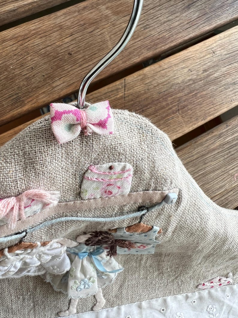 K&ouml;nnte beinhalten: Ein dekorativer, handgefertigter Kleiderb&uuml;gel mit einer leinenartigen Stoffbasis. Er verf&uuml;gt &uuml;ber eine kleine rosa Schleife, Stoffverzierungen und ein skurriles Design eines M&auml;dchens in einem Kleid. Der B&uuml;gel h&auml;ngt an einem silbernen Metallhaken.