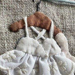 K&ouml;nnte beinhalten: Nahaufnahme eines handgefertigten Textilkunstwerks mit zwei wei&szlig;en Kleidern mit floraler Stickerei, die an einem braunen Kleiderb&uuml;gel h&auml;ngen. Der Hintergrund ist ein strukturierter, neutralfarbiger Stoff mit hellblauen Fadendetails.