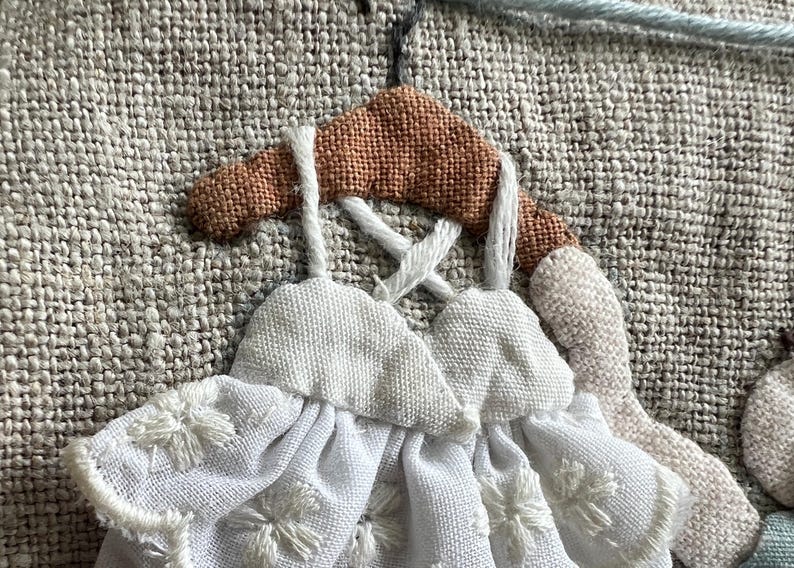 K&ouml;nnte beinhalten: Nahaufnahme eines handgefertigten Textilkunstwerks mit zwei wei&szlig;en Kleidern mit floraler Stickerei, die an einem braunen Kleiderb&uuml;gel h&auml;ngen. Der Hintergrund ist ein strukturierter, neutralfarbiger Stoff mit hellblauen Fadendetails.