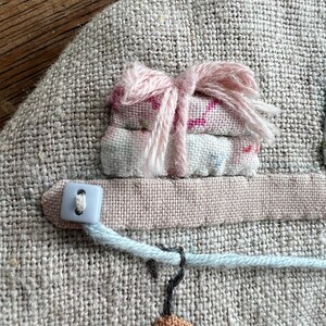 K&ouml;nnte beinhalten: Ein Textilkunstwerk mit einem Miniaturstapel gefalteten Stoffes, der mit rosa Garn gebunden ist. Ein hellblauer Knopf und eine Schnur sind an einem rosa Stoffstreifen befestigt. Unten befindet sich ein kleiner Kleiderb&uuml;gel mit einem braunen und wei&szlig;en Kleidungsst&uuml;ck.