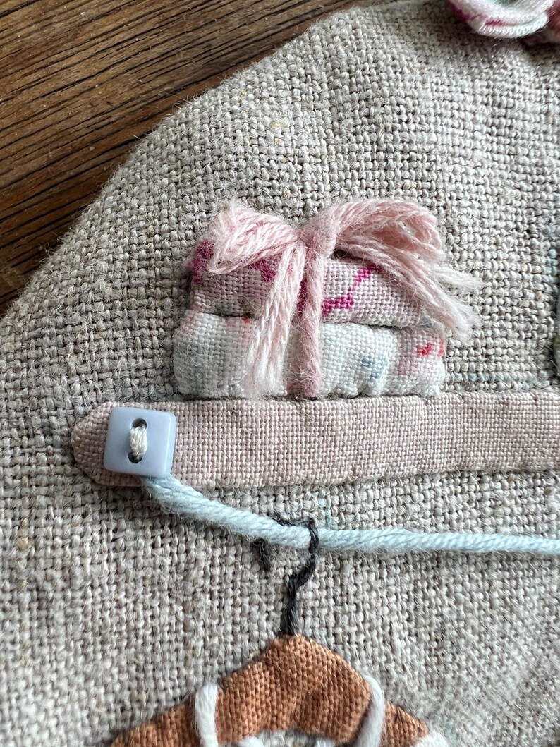 K&ouml;nnte beinhalten: Ein Textilkunstwerk mit einem Miniaturstapel gefalteten Stoffes, der mit rosa Garn gebunden ist. Ein hellblauer Knopf und eine Schnur sind an einem rosa Stoffstreifen befestigt. Unten befindet sich ein kleiner Kleiderb&uuml;gel mit einem braunen und wei&szlig;en Kleidungsst&uuml;ck.