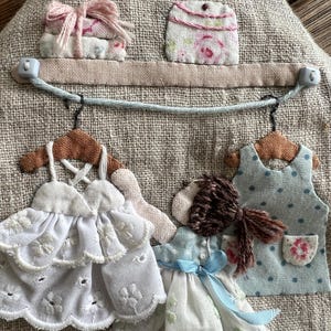 K&ouml;nnte beinhalten: Ein Wandbehang aus Stoff mit Leinenstruktur. Er zeigt Miniatur-Stoffkleider, ein Regal mit Stoffstapeln und eine kleine Geldb&ouml;rse. Die Kleider sind wei&szlig; und blau, mit zarten Stickereien und Spitze. Das Regal ist mit einem rosa Band verziert.