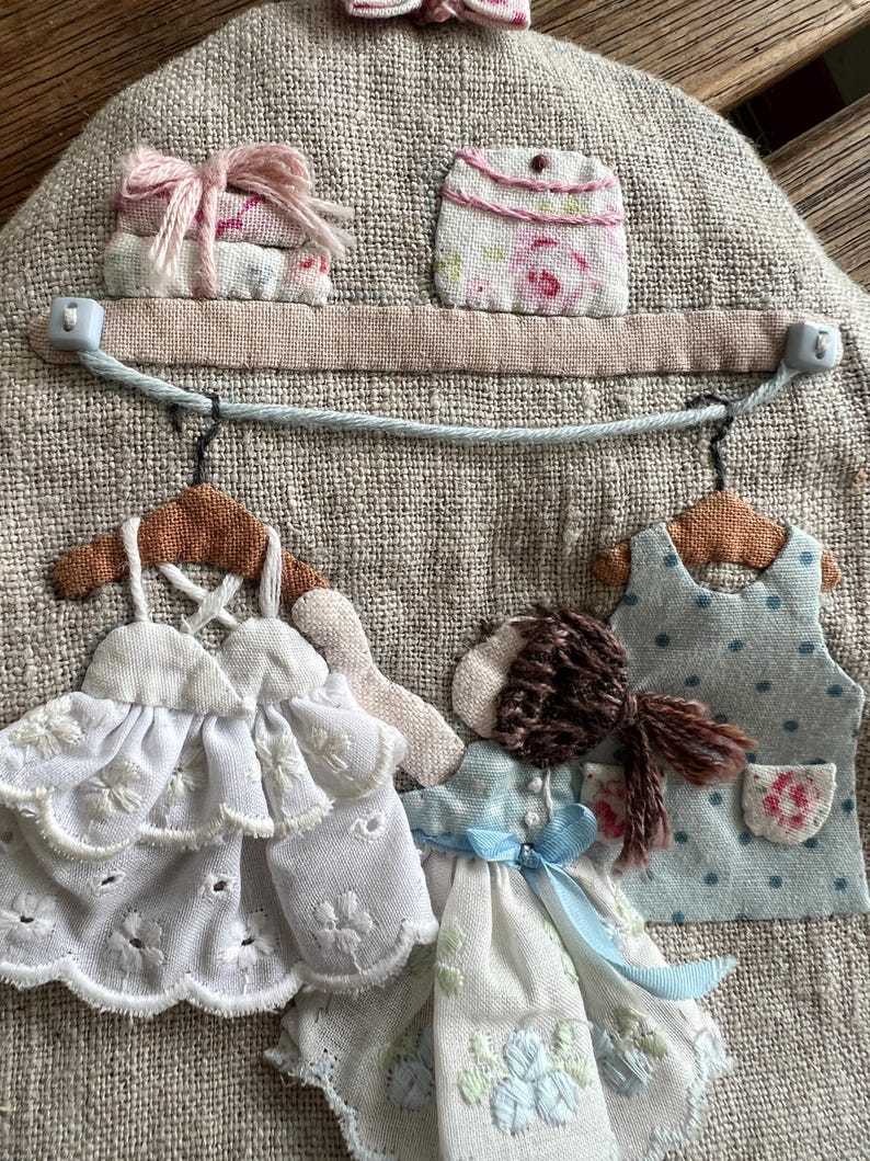 K&ouml;nnte beinhalten: Ein Wandbehang aus Stoff mit Leinenstruktur. Er zeigt Miniatur-Stoffkleider, ein Regal mit Stoffstapeln und eine kleine Geldb&ouml;rse. Die Kleider sind wei&szlig; und blau, mit zarten Stickereien und Spitze. Das Regal ist mit einem rosa Band verziert.