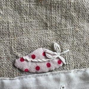 K&ouml;nnte beinhalten: Nahaufnahme eines Leinenstoffs mit einer kleinen, rosa, blattf&ouml;rmigen Applikation. Die Applikation hat rote Tupfen und ist mit wei&szlig;em Faden gebunden. Darunter ein wei&szlig;er Stoff mit gestickten floralen Details.