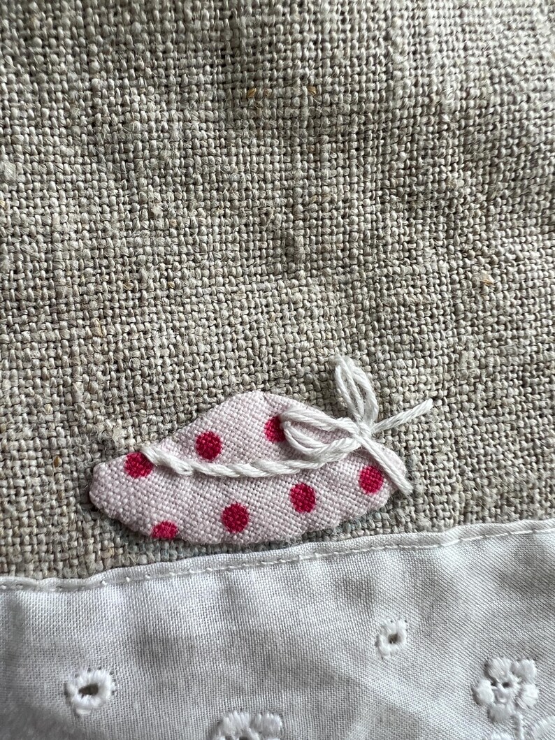 K&ouml;nnte beinhalten: Nahaufnahme eines Leinenstoffs mit einer kleinen, rosa, blattf&ouml;rmigen Applikation. Die Applikation hat rote Tupfen und ist mit wei&szlig;em Faden gebunden. Darunter ein wei&szlig;er Stoff mit gestickten floralen Details.