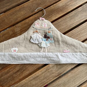 Puede incluir: Una percha decorativa con una base de tela de lino beige, adornada con detalles bordados. Cuenta con un ribete de encaje blanco, una pequeña figura de muñeca y vestidos en miniatura. La percha tiene un gancho de metal y un lazo rosa.