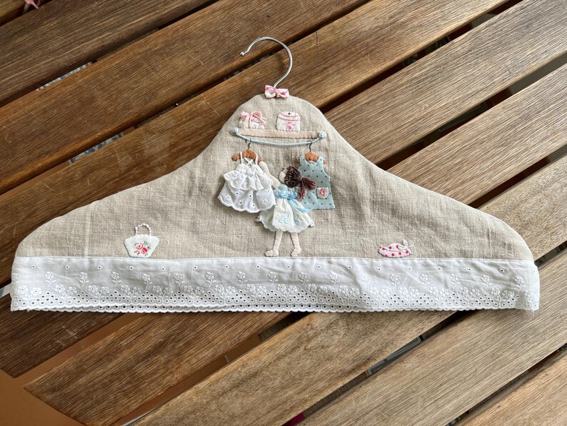 K&ouml;nnte beinhalten: Ein dekorativer Kleiderb&uuml;gel mit einer Basis aus beigem Leinenstoff, verziert mit gestickten Details. Er hat eine wei&szlig;e Spitzentrim, eine kleine Puppenfigur und Miniaturkleider. Der B&uuml;gel hat einen Metallhaken und eine rosa Schleife.
