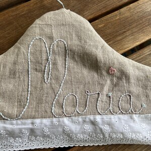 K&ouml;nnte beinhalten: Ein gepolsterter Kleiderb&uuml;gel aus beigem Leinen, bestickt mit dem Namen "Maria" in hellblauer Farbe. Ein kleines rosa Herz ist &uuml;ber dem Namen aufgestickt. Der untere Rand hat eine wei&szlig;e Spitzenschneide mit floralem Muster.