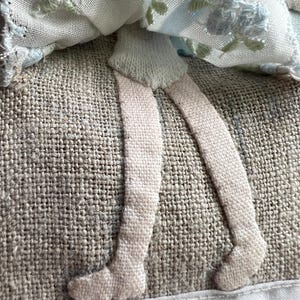 K&ouml;nnte beinhalten: Nahaufnahme einer handgefertigten Textilpuppe mit gestickten floralen Details. Die Beine der Puppe sind aus hellrosa Stoff gefertigt, und das Kleid weist blaue und gr&uuml;ne Blumenstickereien auf. Die Puppe ist vor einem strukturierten, neutralfarbenen Hintergrund platziert.