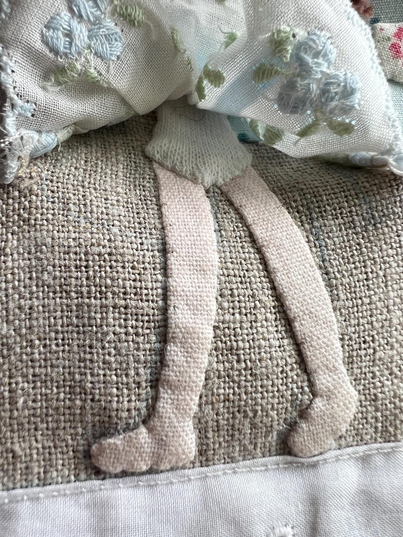 K&ouml;nnte beinhalten: Nahaufnahme einer handgefertigten Textilpuppe mit gestickten floralen Details. Die Beine der Puppe sind aus hellrosa Stoff gefertigt, und das Kleid weist blaue und gr&uuml;ne Blumenstickereien auf. Die Puppe ist vor einem strukturierten, neutralfarbenen Hintergrund platziert.