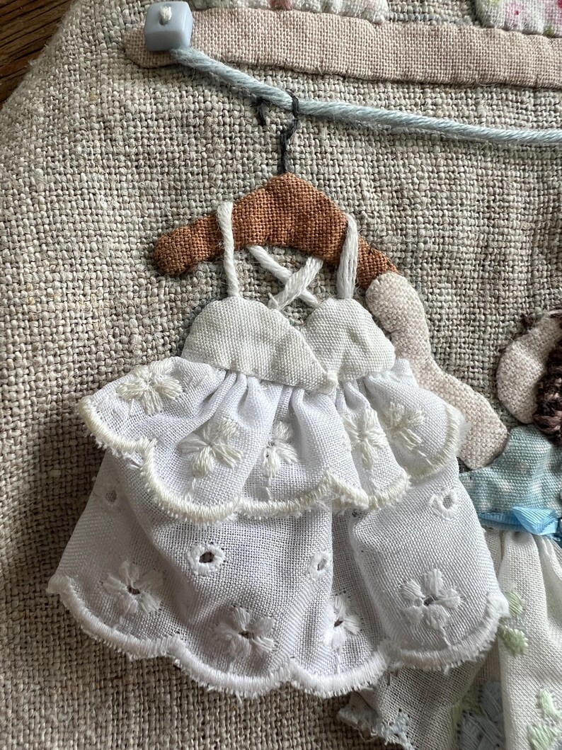 K&ouml;nnte beinhalten: Nahaufnahme eines Textilkunstwerks mit einem Miniatur-Kleidchen in Wei&szlig; mit floraler Stickerei, h&auml;ngend an einem braunen Kleiderb&uuml;gel. Das Kleid hat ein mehrschichtiges Design vor einem strukturierten, neutralfarbenen Hintergrund.